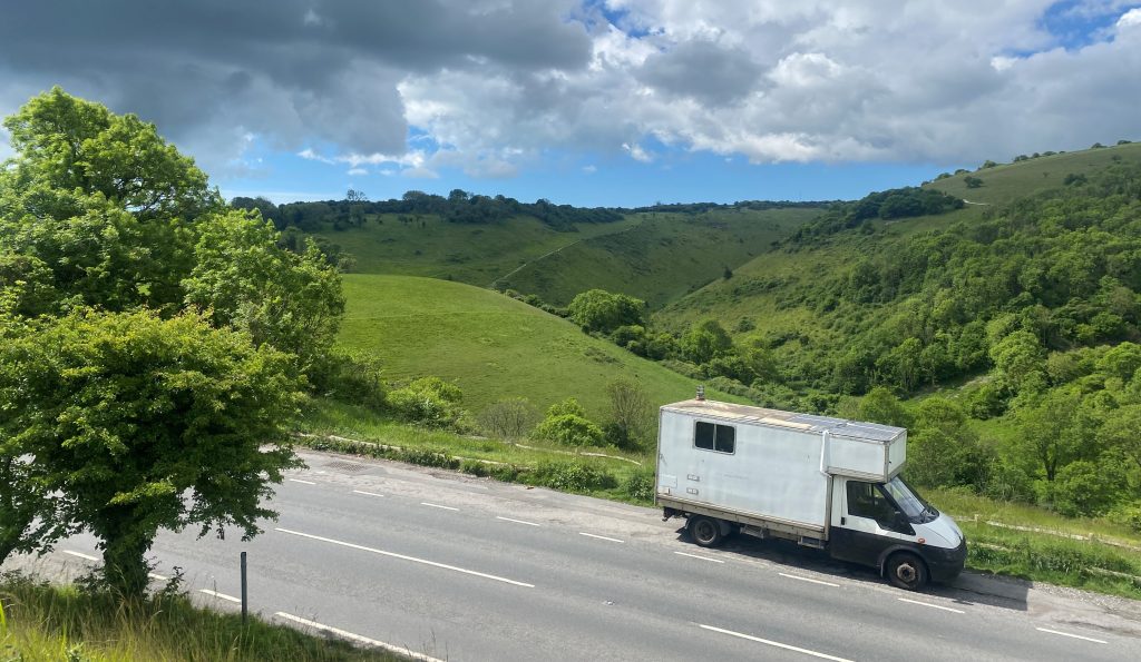 White van driving down a road against a background of green hills and and a tree to the left.