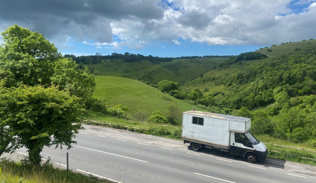 White van driving down a road against a background of green hills and and a tree to the left.