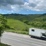 White van driving down a road against a background of green hills and and a tree to the left.