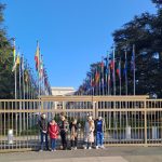 A group of Gypsy, Roma and Traveller children are lined up in front of railings. Behind them the flags of all member states of the United Nations are displayed on the lawn outside the United Nations office.