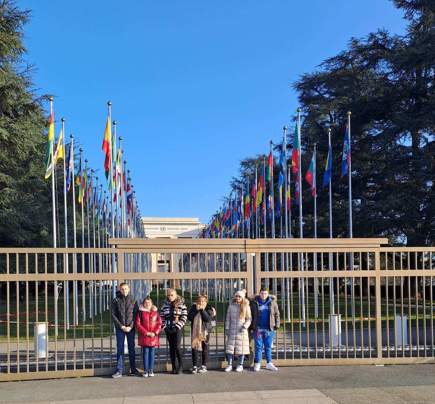 A group of Gypsy, Roma and Traveller children are lined up in front of railings. Behind them the flags of all member states of the United Nations are displayed on the lawn outside the United Nations office.