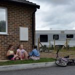A group of three girls are sat on a patch of grass in front of a brick wall with a window. There is a bike laid down on the kerb in front of them and another rested against a small wooden fence to the right, behind them. A caravan can also be seen to the right of the children in the background.