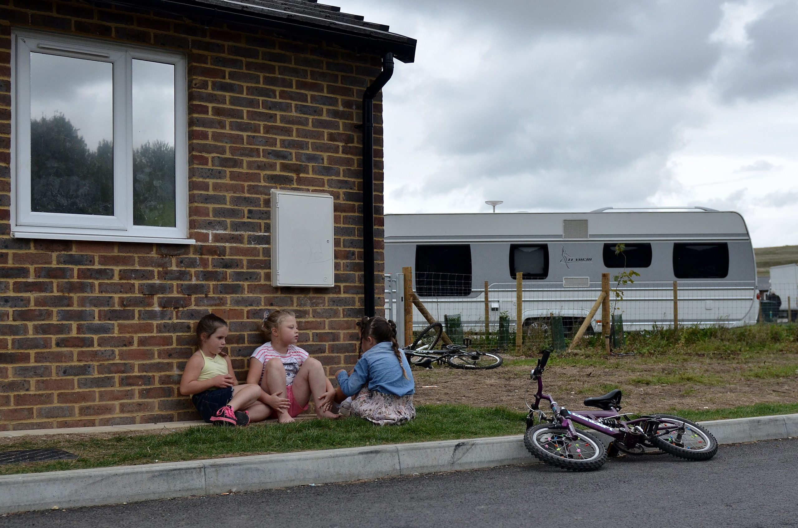 A group of three girls are sat on a patch of grass in front of a brick wall with a window. There is a bike laid down on the kerb in front of them and another rested against a small wooden fence to the right, behind them. A caravan can also be seen to the right of the children in the background.
