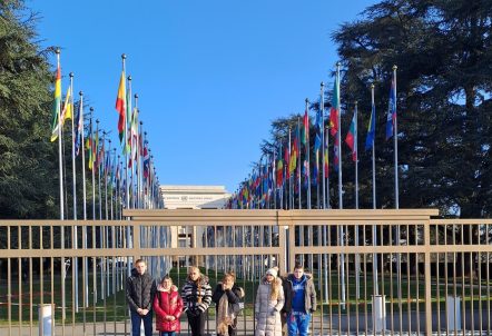 A group of Gypsy, Roma and Traveller children are lined up in front of railings. Behind them the flags of all member states of the United Nations are displayed on the lawn outside the United Nations office.