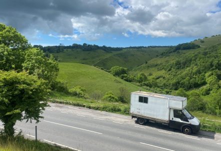 White van driving down a road against a background of green hills and and a tree to the left.