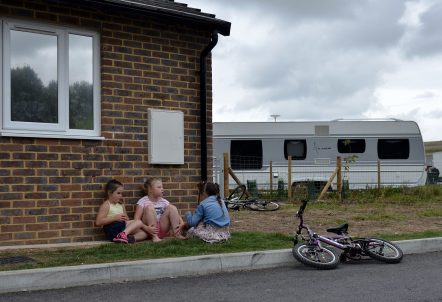 A group of three girls are sat on a patch of grass in front of a brick wall with a window. There is a bike laid down on the kerb in front of them and another rested against a small wooden fence to the right, behind them. A caravan can also be seen to the right of the children in the background.